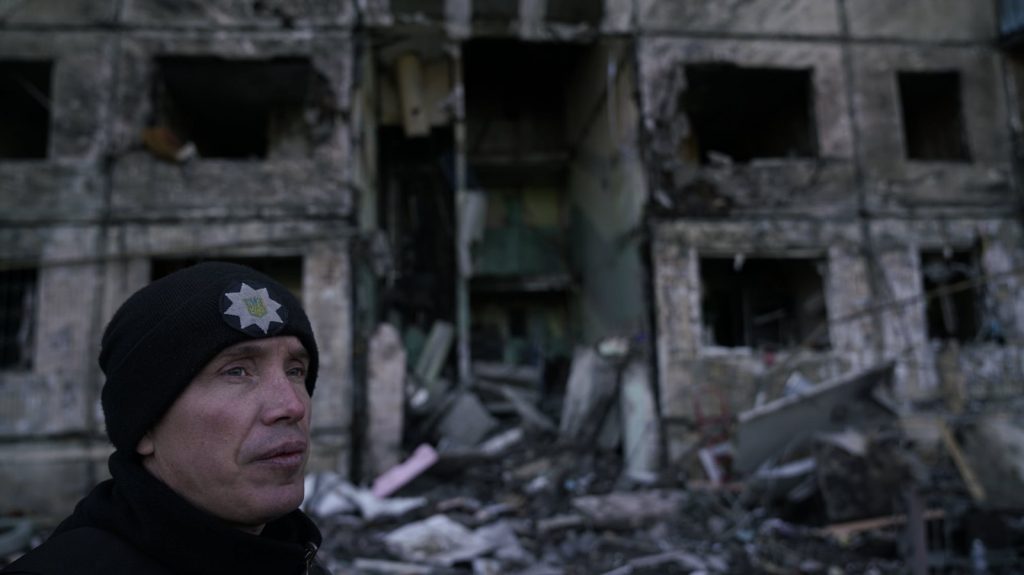 close up of man in wearing black beanie in front of bombed, destroyed building