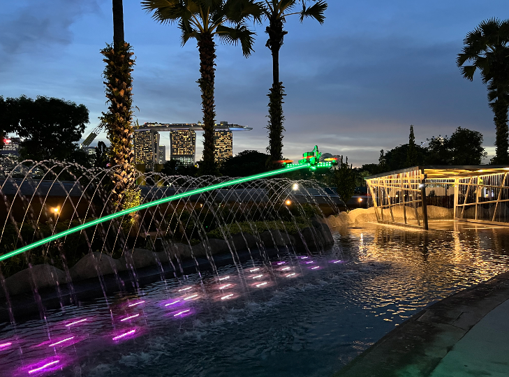 drone racing near fountain over body of water with green trail light and marina bay sands visible in the background