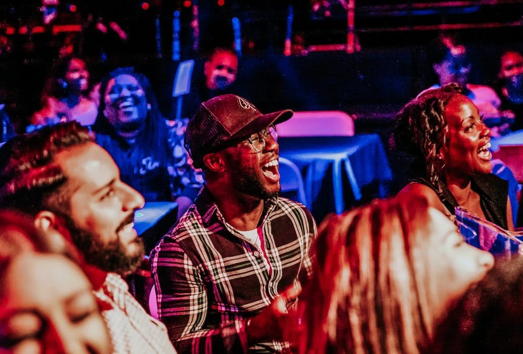 crowd laughing at comedy show, multiple audience members, blue-purple lighting